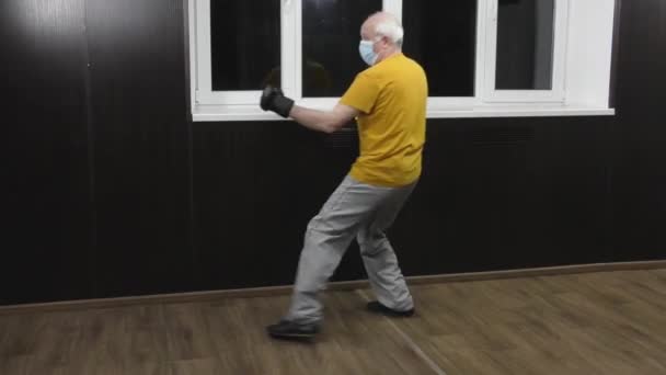 一位戴着医疗面罩的老年运动员 胳膊上戴着黑色垫子 从半跳中训练出一拳 — 图库视频影像