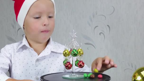 戴着圣诞老人帽的快乐的孩子在一棵小圣诞树旁边吃糖果 — 图库视频影像
