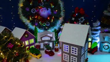 Dekorasyonlu bir evin yakın çekimi. Arka planda büyük bir Noel çelengi ve köknar ağaçları olan büyük bir Noel köyünün önünde dönüyor.