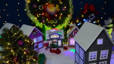 Arka planda büyük bir Noel çelengi ve köknar ağaçları olan büyük bir Noel kasabasının önünde dönen bir evin yakın çekimi.