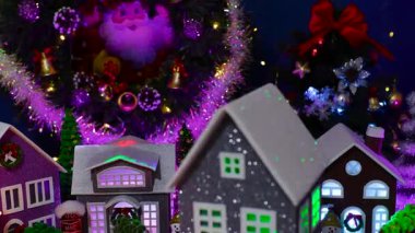 Dekorasyonlu gri bir evin yakın çekimi. Arka planda köknar ağaçları ve büyük bir Noel çelengi olan büyük bir Noel köyünün yakınında dönüyor.