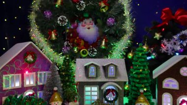 Bir Noel kasabasında büyük bir Noel ağacı ve Noel çelengi karşısında renkli arabalar ve kardan adamlar evlerin önünde duruyor.