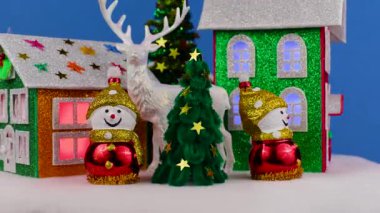 Mavi arka planda, pencerelerinde parlak ışıklar ve küçük yeşil camlı Noel ağaçları ve geyikli kardan adamlar olan dönen evlerin yakın çekimleri.