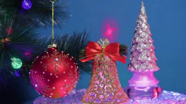 Mavi bir arka planda, kırmızı bir Noel balosunun yanında renkli lazer desenleri olan büyük bir Noel ağacı dalında, gümüş bir Noel ağacı olan altın bir çan.