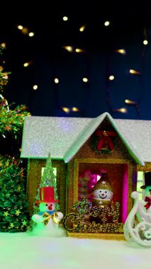 Koyu mavi bir arka planda, yanıp sönen ışıklarla, Noel ağaçlarıyla çevrili bir Noel evi, verandada bir kardan adam ve yakındaki kırmızı ve siyah şapkalı kardan adamlar.