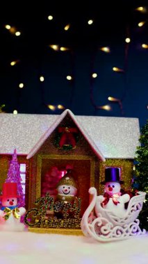 Koyu mavi bir arka planda yanıp sönen bir çelenkle verandada harflerle bir kardan adam ve kırmızı şapkalı bir kardan adam ve yanında beyaz bir kızakla siyah şapkalı bir kardan adam.