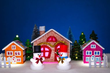 Küçük bir Noel köyünün önünde siyah ve kırmızı şapkalı beyaz kardan adamlar var.