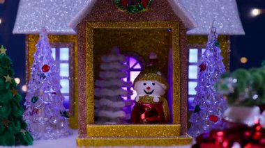 Verandasında bir kardan adam ve Noel ağacı olan bir ev, Noel ağaçları ve bir araba ile çevrili.