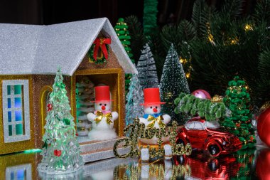 Büyük bir Noel ağacının altında, kırmızı harflerle kızaklı bir kardan adam ve toplarla çevrili kırmızı bir araba ve verandada kırmızı şapkalı bir kardan adam olan bir evin yanındaki Noel ağaçları.