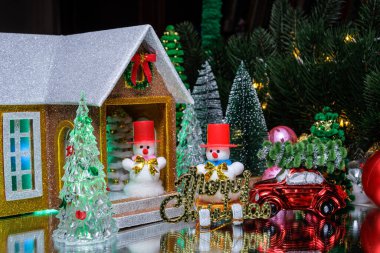 Büyük bir Noel ağacının altında, üzerinde mektuplar olan bir kızak üzerinde bir kardan adam ve çatıda bir Noel ağacı olan kırmızı bir araba, verandada kırmızı şapkalı bir kardan adamın olduğu bir evin yakınında toplar ve Noel ağaçları ile çevrili.