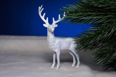 Mavi arka planda, bir geyik heykeli bir Noel ağacının yanında duruyor.