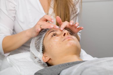 Kozmetik uzmanı ya da cildiyeci güzellik salonunda rahatlamış genç bir bayana yüz masajı yaparken yüzüne krem sürmüş..