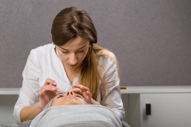 Kozmetik uzmanı ya da cildiyeci güzellik salonunda rahatlamış genç bir bayana yüz masajı yaparken yüzüne krem sürmüş..