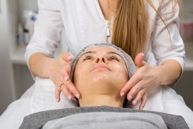 Kozmetik uzmanı ya da cildiyeci güzellik salonunda rahatlamış genç bir bayana yüz masajı yaparken yüzüne krem sürmüş..