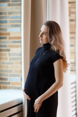 Düşünceli hamile kadın karnını okşuyor ve pencereden dışarı bakıyor..