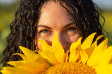 Geniş bir ayçiçeğinin arkasından dışarı bakan genç bir kadının yakın plan portresi. Yaz mevsimi kavramı.