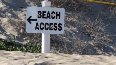 Kumlu bir sahil yolundaki plaj giriş tabelasının el kamerası görüntüsü.