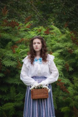 Bahçede elinde hasır bir çantayla eski moda kıyafetler giymiş bir kadın..