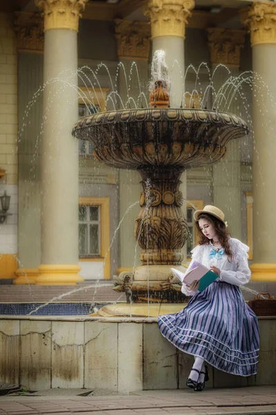 白いドレスの女性は古い建物の背景に噴水に座っている間本を読みます — ストック写真