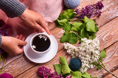 Elinde bir fincan sabah kahvesi ile yukarıdan ahşap arka planda açan bahar leylak çiçekleri dalları var. Dümdüz yerde yatıyordu. Pahalı renkler. Çiçeklerin yaratıcı tasarımı.