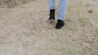 Siyah çizmeli ve kot pantolonlu erkek ayakları kayalık zeminde yürüyor.