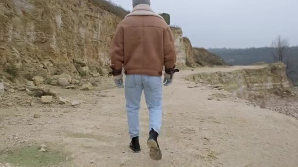 ジャケットとジーンズを着た若い男が捨てられた石のキャリアに沿って歩く — ストック動画
