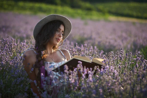 Beautiful girl in hat sitting in purple lavender field and read book.