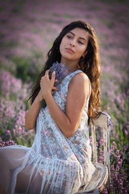 Beyaz sarafanlı güzel bir kız ahşap bir sandalyede oturur ve lavanta tarlasının ortasında poz verir..