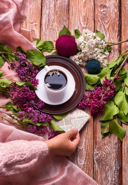 Elinde bir fincan sabah kahvesi ile yukarıdan ahşap arka planda açan bahar leylak çiçekleri dalları var. Dümdüz yerde yatıyordu. Pahalı renkler. Çiçeklerin yaratıcı tasarımı.