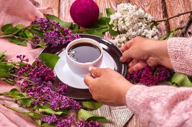 Elinde bir fincan sabah kahvesi ile yukarıdan ahşap arka planda açan bahar leylak çiçekleri dalları var. Dümdüz yerde yatıyordu. Pahalı renkler. Çiçeklerin yaratıcı tasarımı.