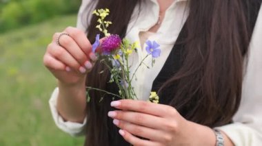 Elinde bir buket kır çiçeğiyle güneşli bir çayırda duran genç bir kadın. Uzun saçlı şapkalı bayan.