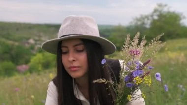 Elinde bir buket kır çiçeğiyle güneşli bir çayırda duran genç bir kadın. Uzun saçlı şapkalı bayan.
