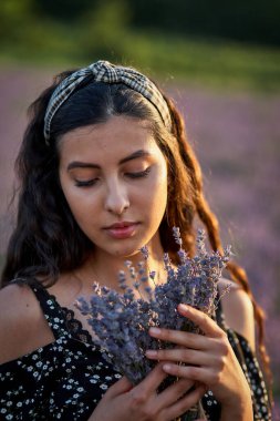 Beyaz elbiseli bir kadının elinde çiçek buketi, yazın lavanta tarlasında çekilmiş bir fotoğrafı.
