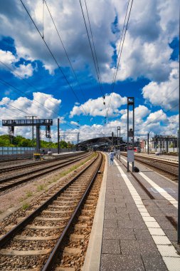 Rail tracks platform under dramatic clouds, expansive commuter hub with overhead catenary, sunlit concrete,