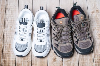 İki çift ayakkabı ahşap zeminde kahverengi yürüyüş ayakkabılarının yanında birbirine zıt ağlarla beyaz spor ayakkabılarını gösteriyor.