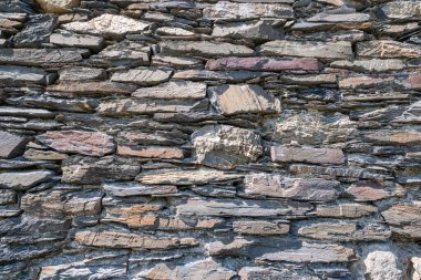 Yıpranmış taş duvar, katmanlı doku, düzensiz istiflenmiş kireç taşı ve granit, görünür havan topu kalıntıları.,