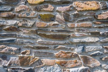 Yıpranmış taş duvar, katmanlı doku, düzensiz istiflenmiş kireç taşı ve granit, görünür havan topu kalıntıları.,