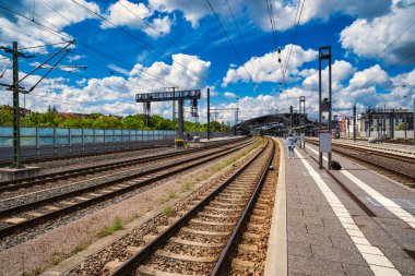 Rail tracks platform under dramatic clouds, expansive commuter hub with overhead catenary, sunlit concrete,