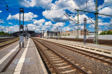 Rail tracks platform under dramatic clouds, expansive commuter hub with overhead catenary, sunlit concrete,