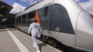 Erfurt, Almanya. 10.05.2025. Beyaz kadın, modern bölgesel taşıma ve koyu turuncu aksanlı, bulutları yansıtan panoramik pencereli, gezgin çeken gümüş tren peronuna çıkıyor.