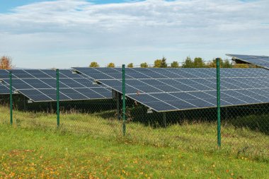 Güneş panelleri mavi gökyüzünün altında dağınık bulutlar, çimenli çayırlarda yere saçılmış raflar, ufuk boyunca sonbahar ağaçları, güneş ışığını yansıtan açılı paneller, ticari