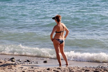 Bikinili ve beysbol şapkalı bir kadın deniz suyunda dikiz aynasından duruyor. Plaj tatili, yüzme ve dinlenme.