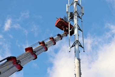 İşçiler mavi gökyüzü ve beyaz bulutların arkasındaki baz istasyonuna anten yerleştiriyorlar. Mobil telekomünikasyon ekipmanları