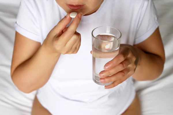 Woman in white top taking pill holding a water glass and sitting on a bed. Concept of taking drugs, sleeping pill, contraception or diet