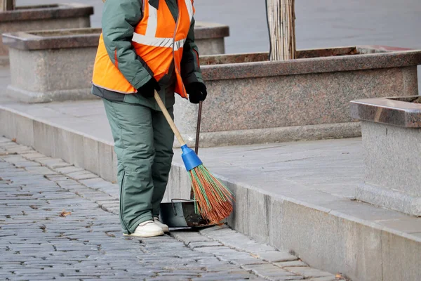 Street cleaning, female municipal worker in uniform sweeping the sidewalk with a broom. Janitor in winter city, concept of unskilled labor