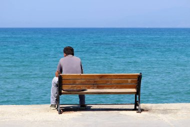 Yalnız bir adam ahşap bir bankta oturuyor mavi deniz arka planında, dikiz manzaralı. Coronavirus salgını sırasında sahilde tatil.