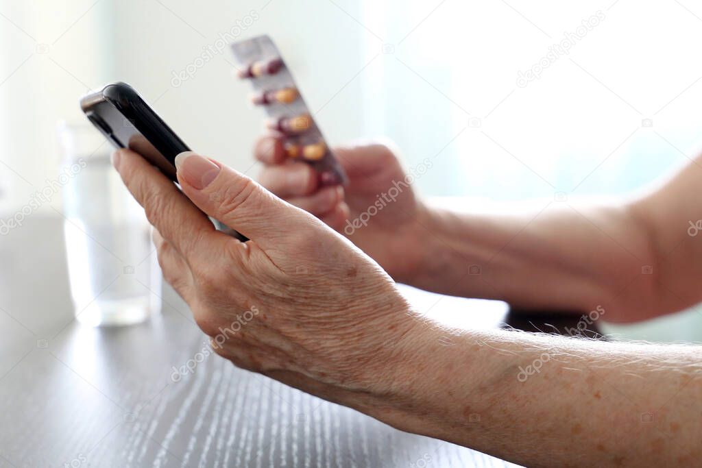 Elderly woman with smartphone sitting at the table, selective focus on wrinkled female hands. Concept of online communication at retirement, search for drug properties, call an ambulance