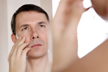 Yakışıklı adam aynada yüzüne dokunan yansımasına bakıyor. Erkek güzelliği, sağlık ve cilt bakımı kavramı