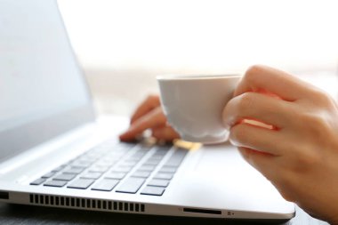 Kadın, pencerenin arkasındaki dizüstü bilgisayarın başında oturup kahve içiyor. Bir bardak sıcak içecek kadın elinde, güneş ışığında, günaydın, iş sırasında mola.