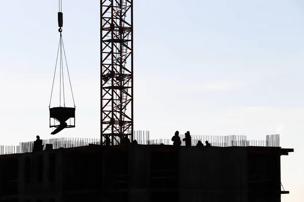 Silhouettes of workers and tower crane with cargo on construction site against the sky. Housing construction, builders working on scaffolding